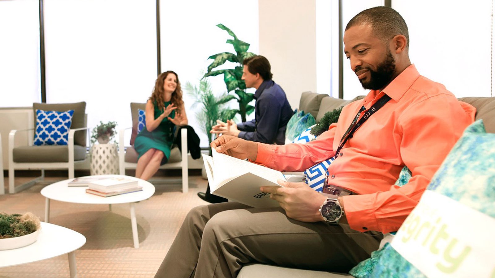 A Rümi employee wearing a bright red shirt smiles as he reads from a folio on a beige couch in the office. In the background, employees smile and collaborate. 