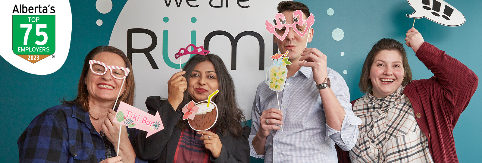 Four Rümi employees smile with party decorations in front of a wall that says "We are Rümi". A logo shows that Rümi is one of Alberta's Top 75 Employers. 