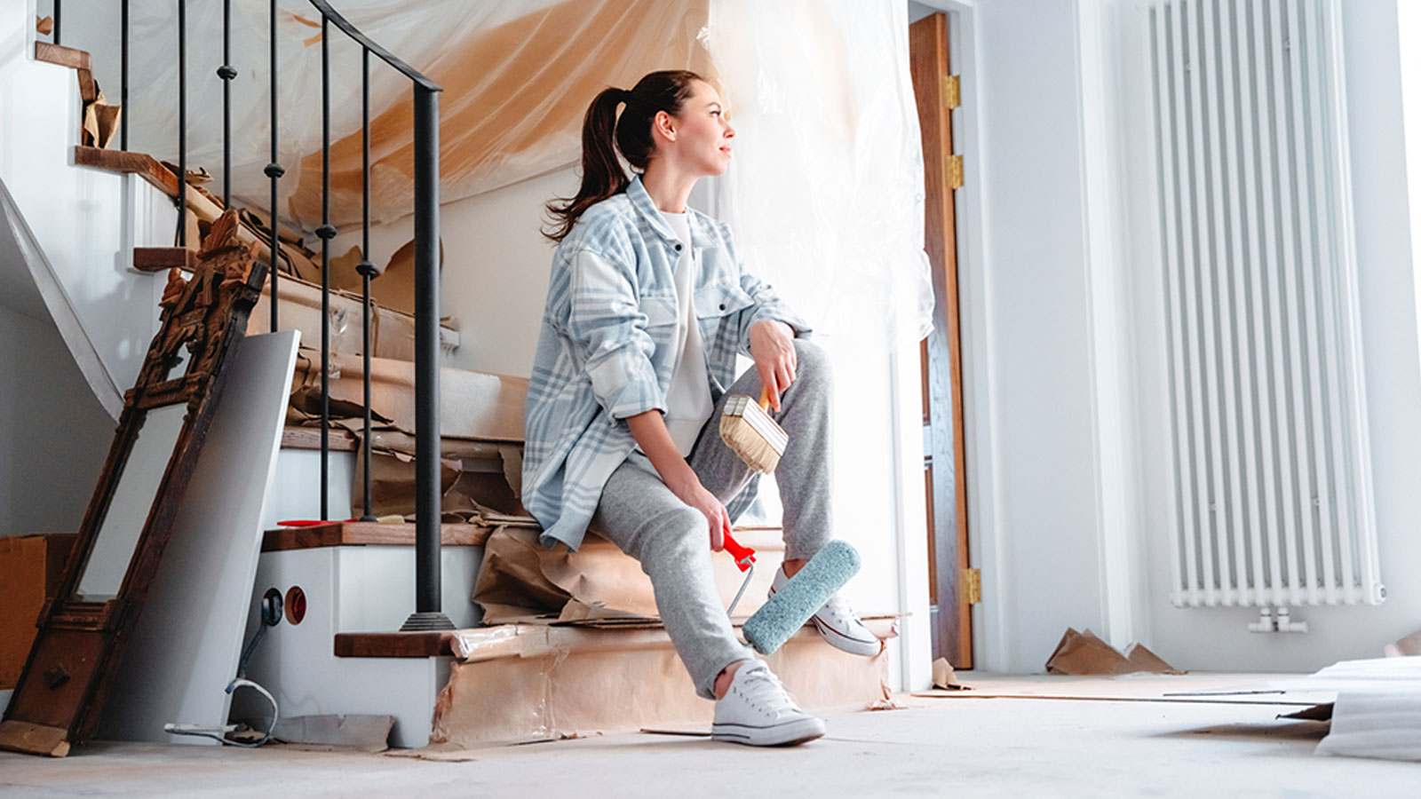A homeowner sits on the stairs while painting her home holding a paintbrush and roller. 