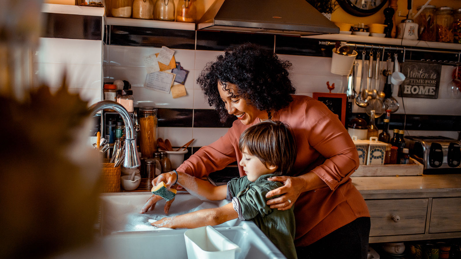 A woman teaches her son to wash dishes with a sponge in the kitchen sink. 