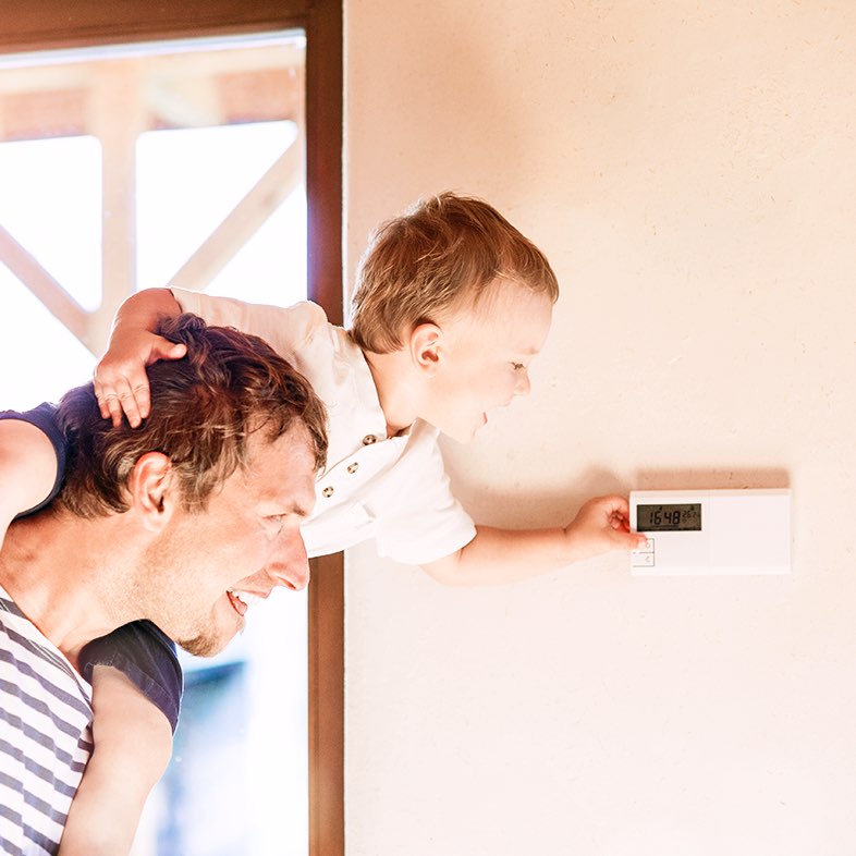 A man holds his young son on his shoulders to reach and adjust the home thermostat. 