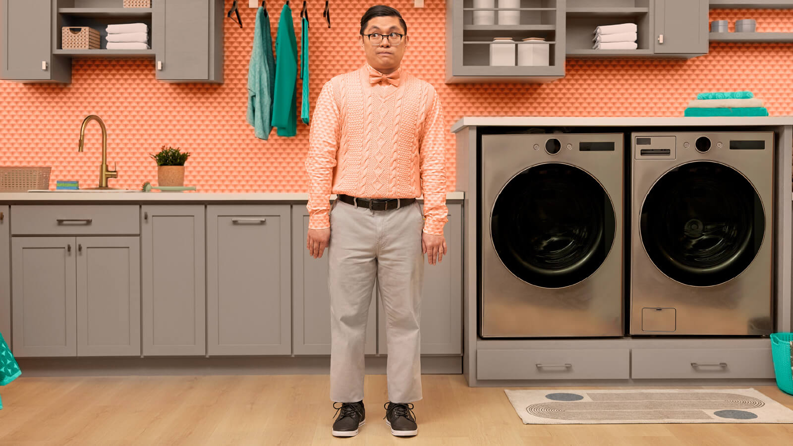 A man wearing a pink sweater vest shrugs looking off to the side in a kitchen with a pink textured wall. 